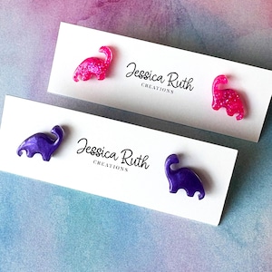 May include: Two pairs of dinosaur-shaped stud earrings displayed on white cards with the text Jessica Ruth Creations. One pair is bright pink with glitter, and the other pair is a solid deep purple. Each earring features a long-necked dinosaur.