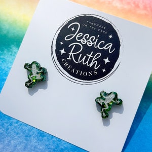 Può includere: Un paio di orecchini a bottone a forma di rana con glitter verde su una carta bianca con il testo "Handmade on the Cape Jessica Ruth Creations".