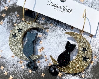 May include: A pair of gold glitter crescent moon earrings with black cat silhouettes and small stars. The earrings are on a white card with the text "Jessica Ruth Creations".