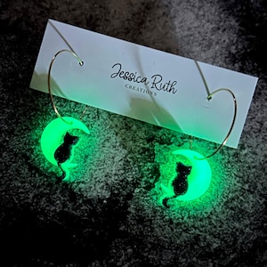 Puede incluir: Pendientes que brillan en la oscuridad con una silueta de gato negro sobre una luna creciente verde. Los pendientes tienen aros dorados. Los pendientes se muestran en una tarjeta blanca con el texto "Jessica Ruth CREATIONS".