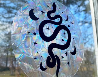 May include: A round iridescent sticker with a black snake design, crescent moons, and stars. The sticker has a holographic finish.