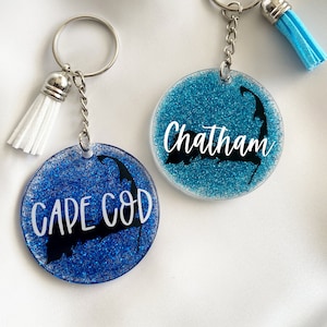 以下が含まれることがあります： 青いグリッターが付いた2つの丸いキーホルダー。1つのキーホルダーには、ケープコッド半島の黒いシルエットの上に白い筆記体で「Cape Cod」と書かれています。もう1つは、白い筆記体で「Chatham」と書かれています。