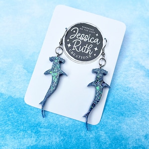 Peut inclure: Une paire de boucles d'oreilles en forme de requin marteau bleu et vert pailleté avec des crochets argentés. Les boucles d'oreilles sont présentées sur une carte blanche avec le texte "Handmade on the Cape Jessica Ruth Creations".