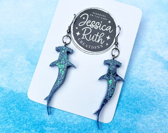 May include: A pair of blue and green glitter hammerhead shark earrings with silver hooks. The earrings are displayed on a white card with the text "Handmade on the Cape Jessica Ruth Creations".