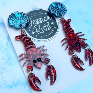 May include: Red glitter lobster earrings with blue glitter seashell charms. The earrings are handmade by Jessica Ruth Creations.