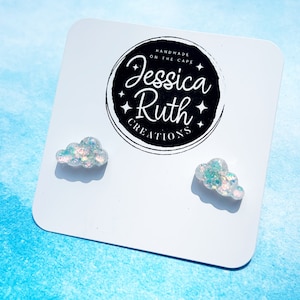 May include: A pair of white cloud-shaped earrings with iridescent glitter. The earrings are on a white card with the text "Handmade on the Cape Jessica Ruth Creations".