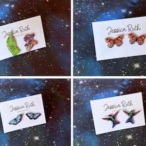 May include: Four sets of stud earrings on white display cards labelled Jessica Ruth Creations. Designs include a green leaf and mushroom, orange butterflies, blue butterflies, and hummingbirds. Each pair features detailed, colourful illustrations.