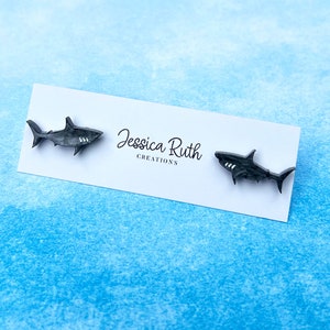 Pode incluir: Um par de brincos de tubarão pretos em um cartão branco com o texto "Jessica Ruth Creations".
