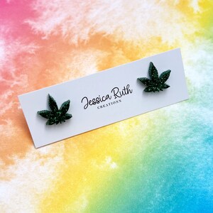 May include: A pair of dark green glittery cannabis leaf-shaped stud earrings attached to a white rectangular display card. The card features the black printed text Jessica Ruth CREATIONS centered between the two earrings.