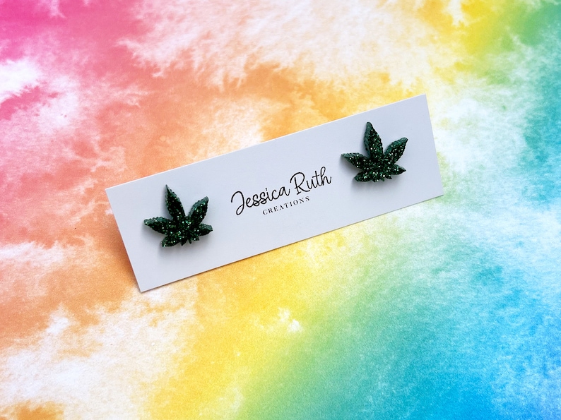 May include: A pair of dark green glittery cannabis leaf-shaped stud earrings attached to a white rectangular display card. The card features the black printed text Jessica Ruth CREATIONS centered between the two earrings.