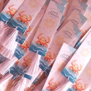May include: A collection of personalized bookmarks featuring a cartoon crab on a blue and white background. Each bookmark has a tassel and the name "Lons Camillo" printed on it. The bookmarks are likely for a baptism or christening.