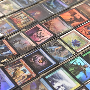 May include: A collection of Magic: The Gathering trading cards, displayed in protective sleeves. The cards feature various fantasy art illustrations, with a range of colors and designs. The cards are arranged in a grid-like pattern.