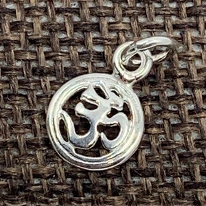 May include: A small, silver-colored pendant with the Om symbol, a sacred sound and spiritual icon in Hinduism, Buddhism, and Jainism. The pendant is circular with a loop for a chain. The background is a textured, brown fabric.