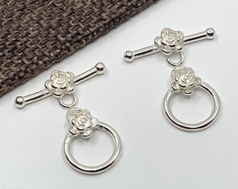 Op de afbeelding: Twee zilveren toggle-sluitingen met een rozenontwerp. Elke sluiting heeft een kleine ring en een staaf met een bal aan elk uiteinde.