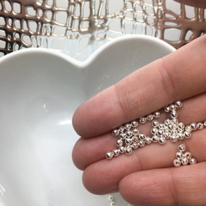 May include: A close-up of a hand holding a pile of small, silver beads. The beads are round and smooth, and they are scattered in a random pattern.