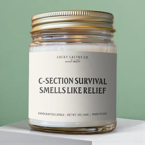 May include: A clear glass jar candle with a gold lid. The label reads "C-SECTION SURVIVAL SMELLS LIKE RELIEF" and "LUCKY CACTUS CO." The candle is handcrafted and weighs 7 oz (198g).