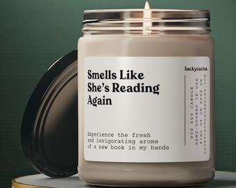 May include: A 9oz soy candle in a clear glass jar with a black lid. The white label features the black text Smells Like She’s Reading Again, Experience the fresh and invigorating aroma of a new book in my hands, and luckycactus branding.