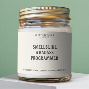 May include: A glass jar candle with a gold lid. The label reads "SMELLS LIKE A BADASS PROGRAMMER" in bold white letters. The candle is handcrafted and weighs 7 oz (198g). The brand name is Lucky Cactus Co.