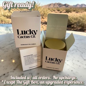 a gift box for a cactus candle from Lucky Cactus Co., with a desert landscape in the background. The gift box is open, revealing a gold-colored candle inside. The text on the gift box indicates that it is included in all orders, with no upcharge, and that it is an upgraded experience compared to the gift box.