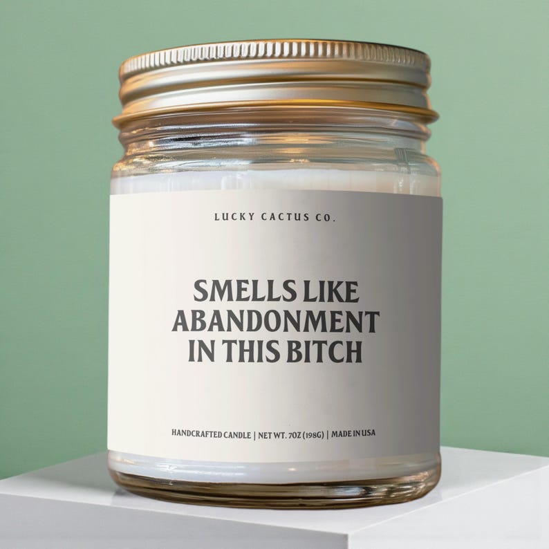 May include: A clear glass jar candle with a gold lid. The label reads "LUCKY CACTUS CO." and "SMELLS LIKE ABANDONMENT IN THIS BITCH." The candle is handcrafted and weighs 7 oz (198g).