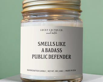 May include: A clear glass candle jar with a gold metal lid, containing white wax. The label reads Lucky Cactus Co. small batch, Smells Like A Badass Public Defender, Handcrafted Candle, Net Wt. 7oz (198g), Made in USA in black text.