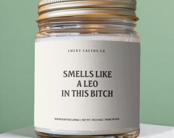 May include: A clear glass candle jar with a gold metal lid and a white label. The label features the text LUCKY CACTUS CO. and SMELLS LIKE A LEO IN THIS BITCH. The bottom of the label states HANDCRAFTED CANDLE | NET WT. 7OZ (198G) | MADE IN USA.