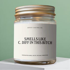 May include: A glass jar candle with a gold lid. The label reads "LUCKY CACTUS CO." and "SMELLS LIKE C. DIFF IN THIS BITCH." The candle is 198g and made in the USA. The candle sits on a white surface.