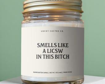 May include: A clear glass candle jar with a gold metal lid, filled with white wax. The label reads LUCKY CACTUS CO. and SMELLS LIKE A LICSW IN THIS BITCH. The bottom of the label states HANDCRAFTED CANDLE | NET WT. 7OZ (198G) | MADE IN USA.