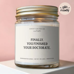 May include: A clear glass candle jar with a gold metal lid and a white label. The label reads Lucky Cactus Co. small batch, FINALLY. YOU FINISHED YOUR DOCTORATE. HANDCRAFTED CANDLE | NET WT. 7OZ (198G) | MADE IN USA. A circular sticker says gift ready.