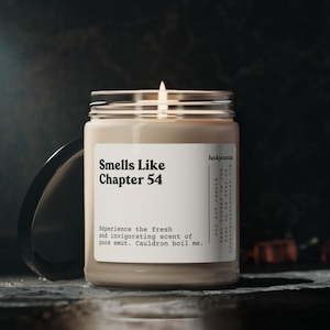 May include: A lit candle in a glass jar with a black lid. The label reads "Smells Like Chapter 54" with additional text. The candle is on a dark surface, with a dark background.