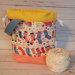 May include: A drawstring bag with a yellow top, a peach bottom, and a blue strap. The bag is printed with a pattern of colorful socks hanging on a clothesline with hearts. A ball of white yarn with multi-colored flecks is in the foreground. The text "The Knitting Owl" is on the bag.