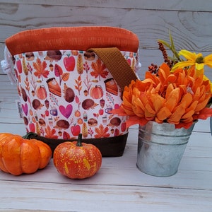May include: A fabric tote bag with a brown base and handle, featuring a fall-themed pattern of leaves, pumpkins, hedgehogs, and coffee cups. The top rim is orange with the words "AUTUMN LEAVES" printed on it. Two small pumpkins and a metal bucket with orange flowers are also in the image.