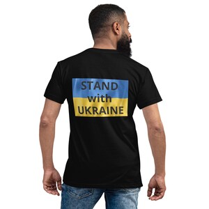 Könnte beinhalten: Schwarzes T-Shirt mit dem Text "STAND with UKRAINE" in weißen Buchstaben über einem blau-gelben ukrainischen Flaggen-Design. Das Shirt hat einen Rundhalsausschnitt und kurze Ärmel.