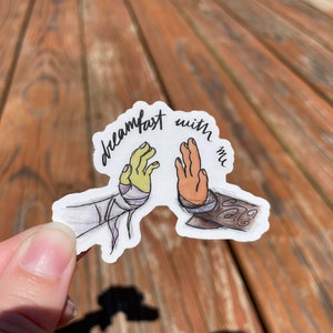 May include: A white sticker with a hand drawn illustration of two hands high-fiving. The text "dreamfast with me" is written in black cursive above the hands.