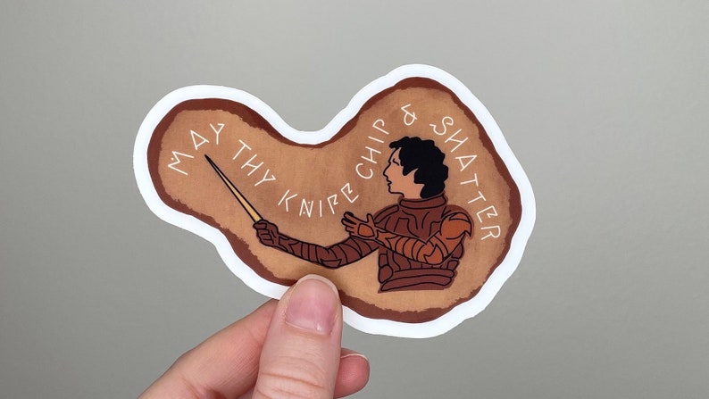 May include: A brown and white sticker with a drawing of a person in armor holding a knife. The text on the sticker reads "May thy knife chip & shatter".