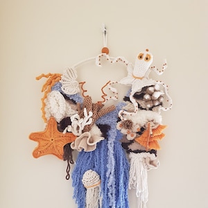 Könnte beinhalten: Ein handgefertigter Wandbehang mit einer gehäkelten Meereslandschaft. Das Kunstwerk umfasst einen Oktopus, Seestern, Korallen und Quallen in Weiß-, Orange-, Braun- und Blautönen. Das Stück ist auf einem weißen Reifen montiert und zum Aufhängen an der Wand bestimmt.