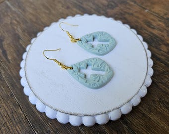 Puede incluir: Un par de pendientes de lágrima azul claro con un recorte en forma de cruz y detalles florales ornamentados. Los pendientes tienen ganchos dorados. Los pendientes se exhiben en una bandeja redonda de madera blanca con borde festoneado.