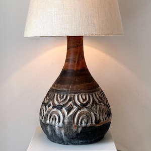 Może przedstawiać: Lampka nocna w stylu vintage z beżowym lnianym abażurem i ciemnobrązową ceramiczną podstawą. Podstawa posiada dekoracyjny wzór w kolorze białym i czarnym. Lampa stoi na białym cokole.