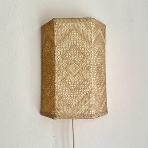 Può includere: Una lampada da parete con un paralume rettangolare testurizzato realizzato in materiale intrecciato color marrone chiaro con un motivo geometrico a rombi ripetuto. Il paralume ha una struttura rigida e un cavo elettrico chiaro sul fondo.