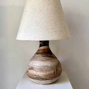 May include: A table lamp featuring a rounded, bulbous ceramic base with horizontal, swirling bands of brown, tan, and cream glaze. The lamp is topped with a tapered, conical lampshade made of a textured, light beige fabric with a visible weave.