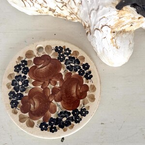 May include: A round, hand-painted ceramic trivet with a floral design. The trivet features three large brown roses surrounded by small black flowers and green leaves on a cream background. A white sheep sculpture is in the background.