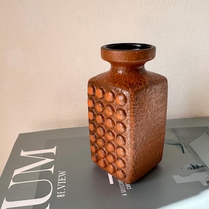 Puede incluir: Un jarrón de cerámica marrón con cuerpo cuadrado y superficie texturizada. El jarrón tiene una serie de patrones circulares en relieve en la parte delantera y los lados. La parte superior del jarrón es negra. El jarrón está sobre una revista.