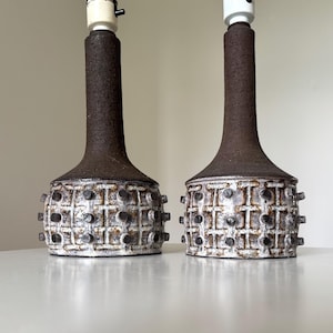 Może przedstawiać: Para ceramicznych lamp stołowych. Każda lampa ma teksturowaną, ciemnobrązową szyję i białą podstawę z wzorem kratki i małymi, ciemnobrązowymi wypukłościami. Lampy mają białe oprawki.