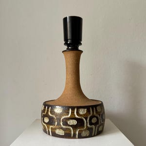 Könnte beinhalten: Eine Tischlampe aus Keramik mit einem braunen, strukturierten Sockel und einem schwarzen Oberteil. Der Sockel weist ein sich wiederholendes Muster aus braunen und cremefarbenen Formen auf. Die Lampe steht auf einer weißen Oberfläche vor einem schlichten Hintergrund. Die Lampe ist etwa 30 cm hoch.