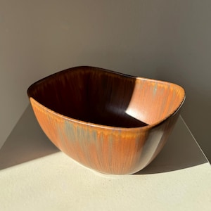 Può includere: Una ciotola rettangolare in ceramica con bordo curvo. La ciotola ha una finitura lucida e screziata nei toni del marrone e dell'arancione. L'interno è marrone più scuro. La ciotola è vuota e poggia su una superficie bianca.
