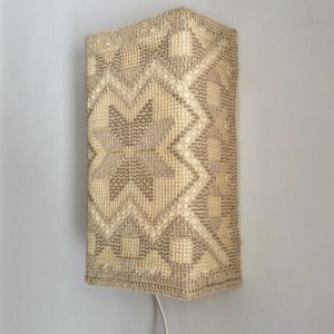 Może przedstawiać: Prostokątny kinkiet ścienny z tkanym, geometrycznym wzorem w odcieniach beżu i kremu. Lampa jest zamontowana na jasnoszarej ścianie i ma biały przewód elektryczny.