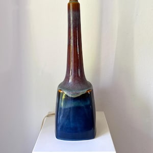 Może przedstawiać: Ceramiczna lampa z ciemnoniebieską podstawą, przechodzącą w czerwonobrązowy korpus, zwieńczona białym uchwytem na żarówkę. Lampa ma unikalny, wydłużony kształt i błyszczące wykończenie, odpowiednie do dekoracji wnętrz.