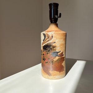 Puede incluir: Una lámpara de mesa de cerámica con un diseño abstracto en marrón, negro y blanco. La lámpara tiene un casquillo negro y un cable.