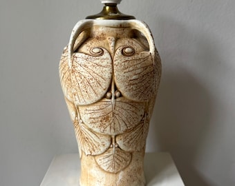 Peut inclure: Un pied de lampe de table en céramique avec un corps couleur crème orné d'un motif de feuilles en relief de couleur brune. La lampe possède deux anses latérales, un col en métal doré et une douille cylindrique blanche.