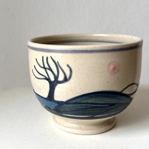 Puede incluir: Un cuenco de cerámica con acabado beige moteado. El cuenco presenta un diseño de árbol y paisaje azul y negro. Hay un punto rosa en el lateral. El borde tiene una franja azul. El cuenco se apoya en un pequeño pie.